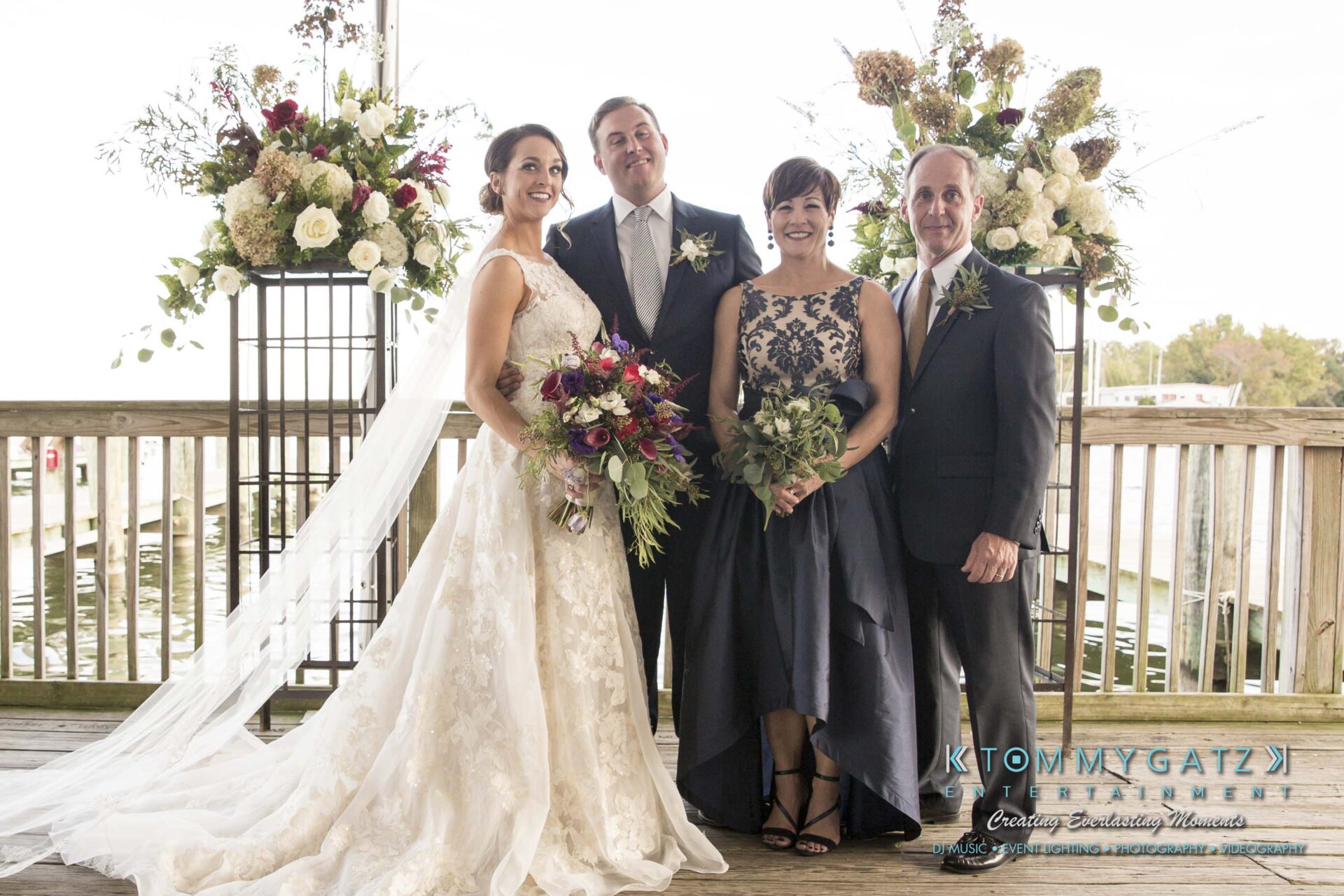 A bride and groom stand with two older adults in front of floral arrangements by a waterfront.