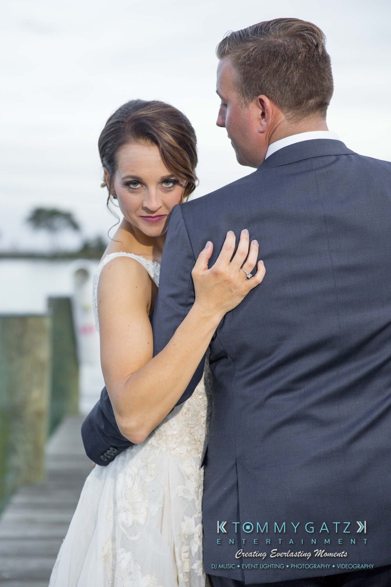 Amanda-Tyler-102117-FORMALS-Low-Res-WM-51 - TOMMY GATZ ENTERTAINMENT A couple in formal attire on a wooden dock, with a lake and trees in the background. The woman in a white dress embraces the man in a suit from behind.