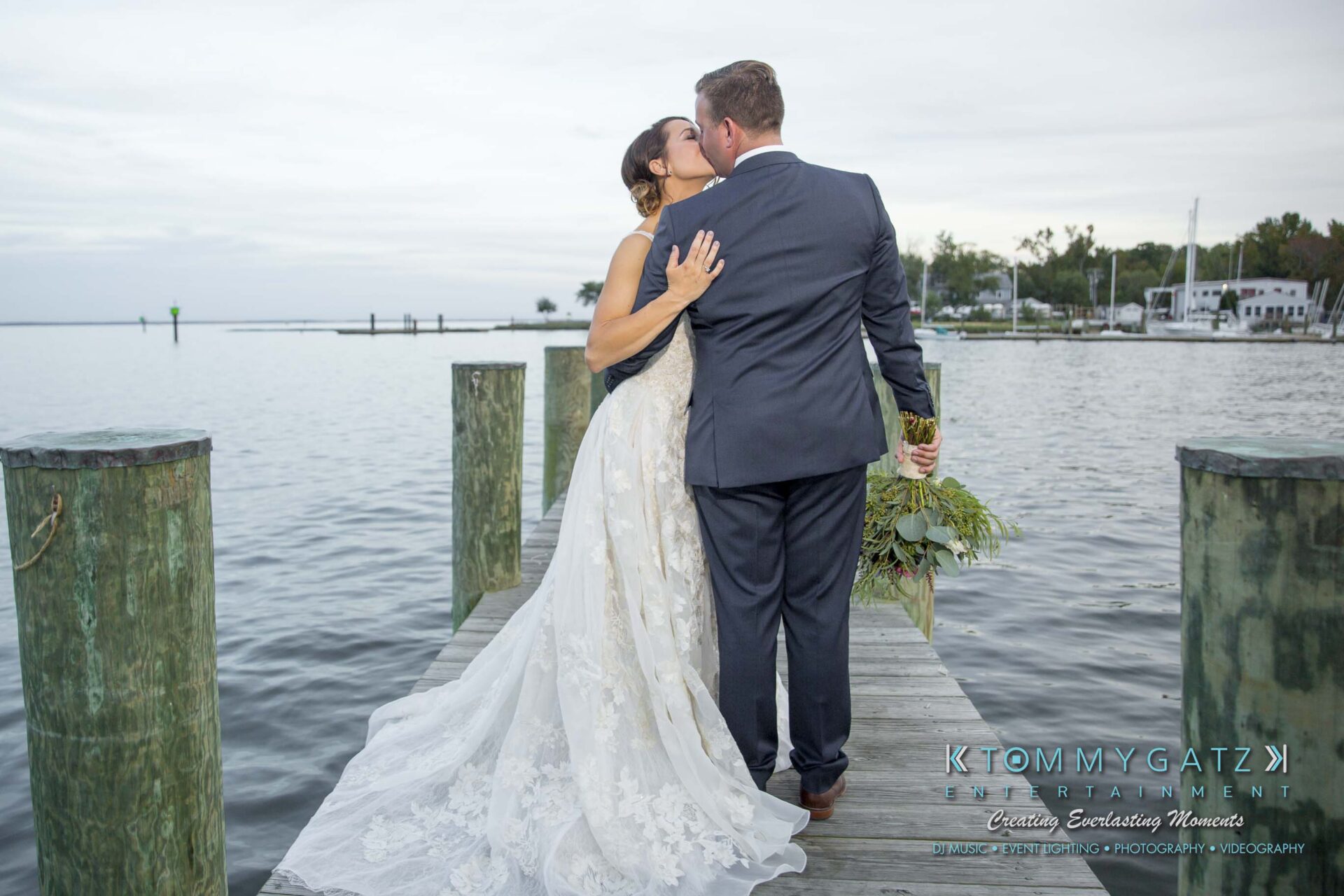 Amanda-Tyler-102117-FORMALS-Low-Res-WM-52 - TOMMY GATZ ENTERTAINMENT A bride and groom kiss on a wooden dock by a calm lake, with trees and a cloudy sky in the background. The bride holds a bouquet.