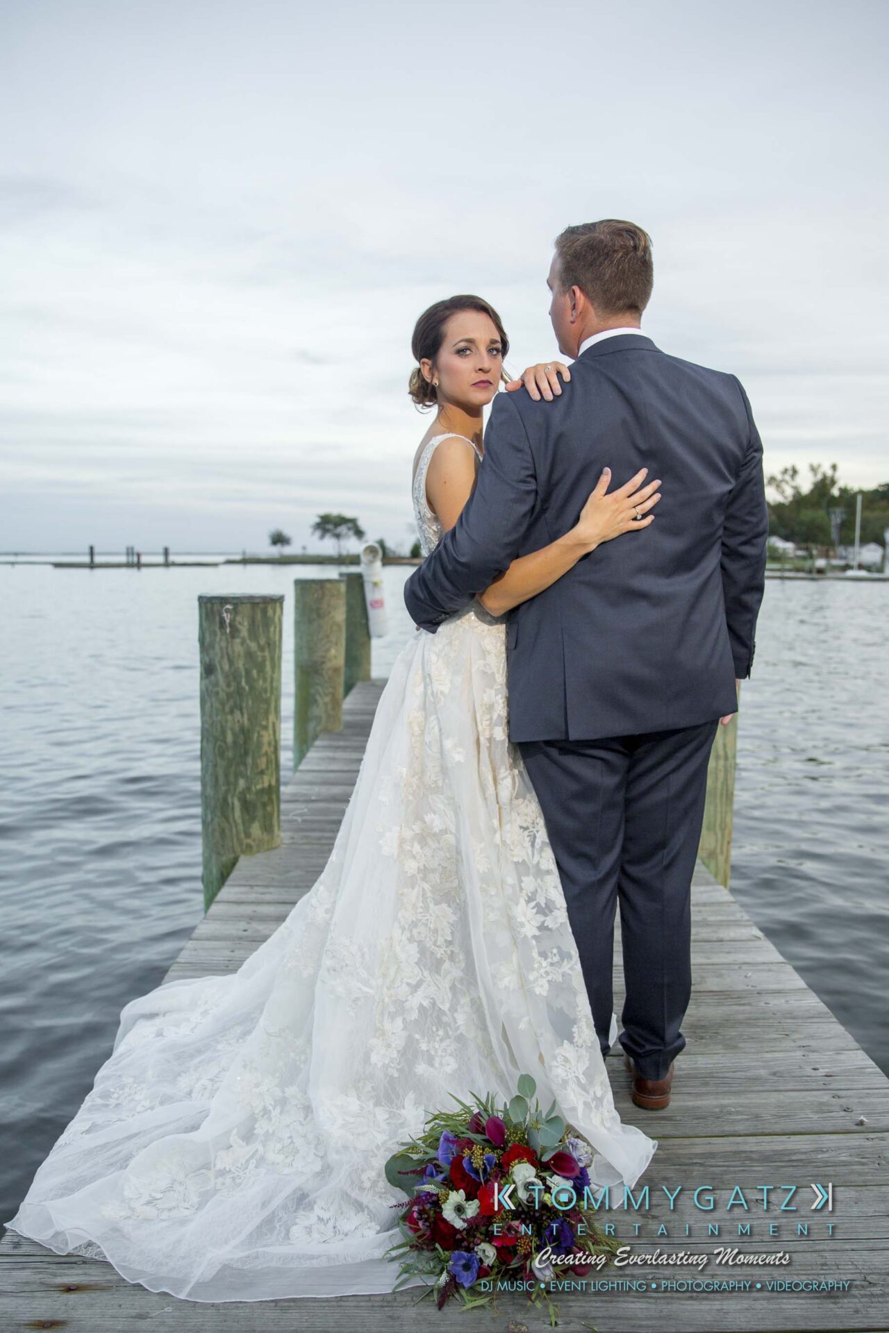 Amanda-Tyler-102117-FORMALS-Low-Res-WM-54 - TOMMY GATZ ENTERTAINMENT A bride and groom stand on a dock by the water, with the bride in a lace dress holding the groom's shoulder. A bouquet lies on the dock.