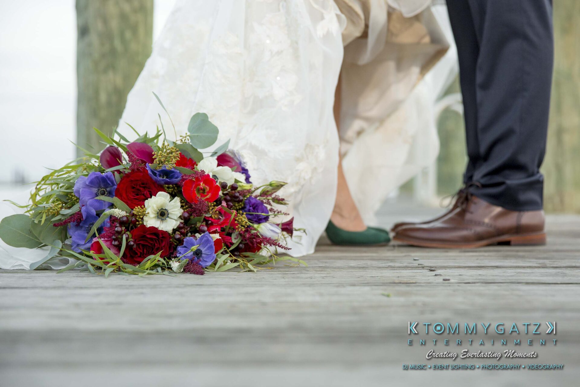 Amanda-Tyler-102117-FORMALS-Low-Res-WM-58 - TOMMY GATZ ENTERTAINMENT A bouquet of colorful flowers lies on a wooden deck near the feet of a bride in a white dress and a groom in a suit.