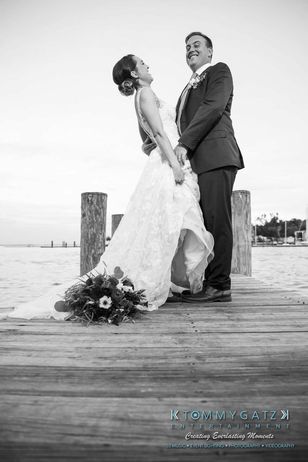 Amanda-Tyler-102117-FORMALS-Low-Res-WM-59 - TOMMY GATZ ENTERTAINMENT Bride and groom share a joyful moment on a wooden dock by the water, with a bouquet on the ground.