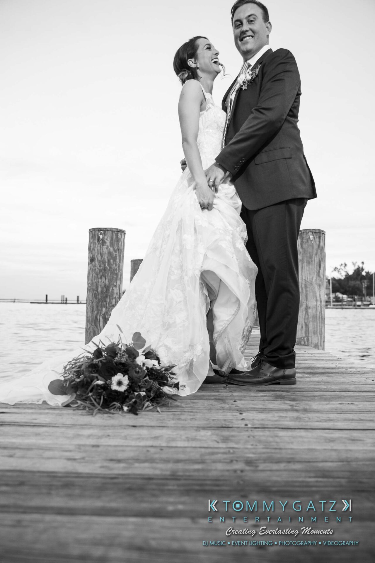 Amanda-Tyler-102117-FORMALS-Low-Res-WM-60 - TOMMY GATZ ENTERTAINMENT Bride and groom smiling on a wooden pier by the water, with a bouquet lying on the ground.