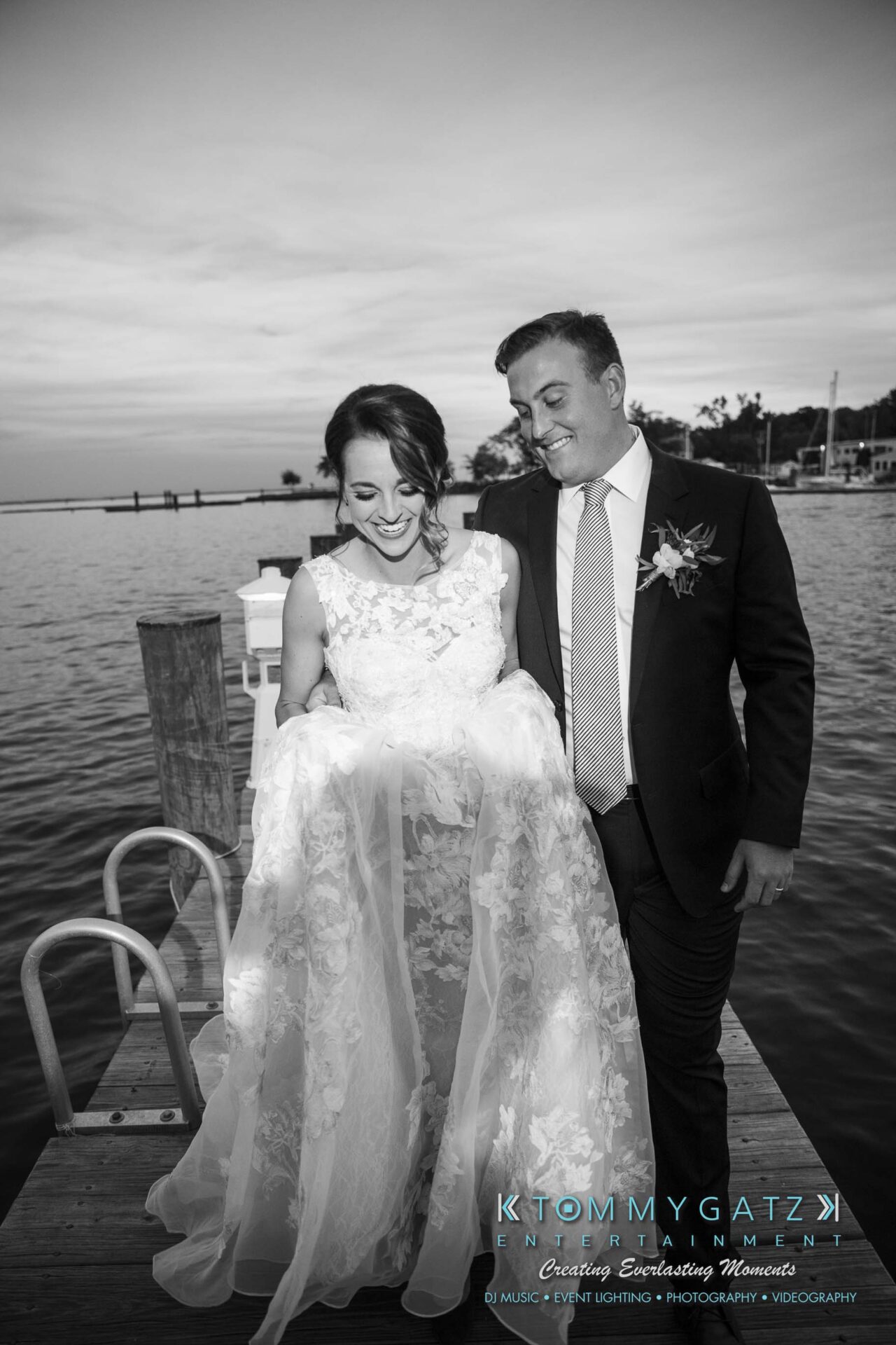 Amanda-Tyler-102117-FORMALS-Low-Res-WM-66 - TOMMY GATZ ENTERTAINMENT A bride and groom smile while standing on a dock by the water, the bride holding her lace dress.
