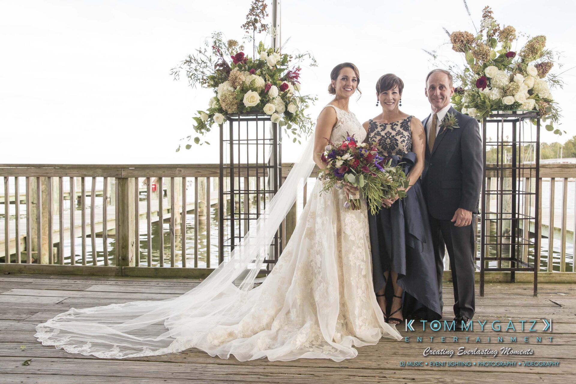 Amanda-Tyler-102117-FORMALS-Low-Res-WM-7 - TOMMY GATZ ENTERTAINMENT Bride in a white dress poses with two people on a wooden deck. A floral arrangement is on one side, and water is visible in the background.
