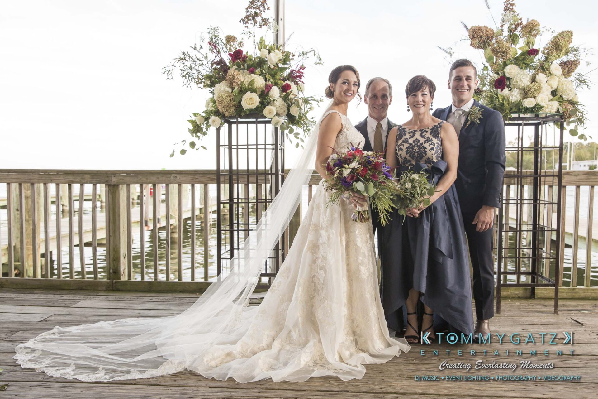 Amanda-Tyler-102117-FORMALS-Low-Res-WM-9 - TOMMY GATZ ENTERTAINMENT A bride and groom stand with two older adults on a wooden deck, with floral arrangements in the background.