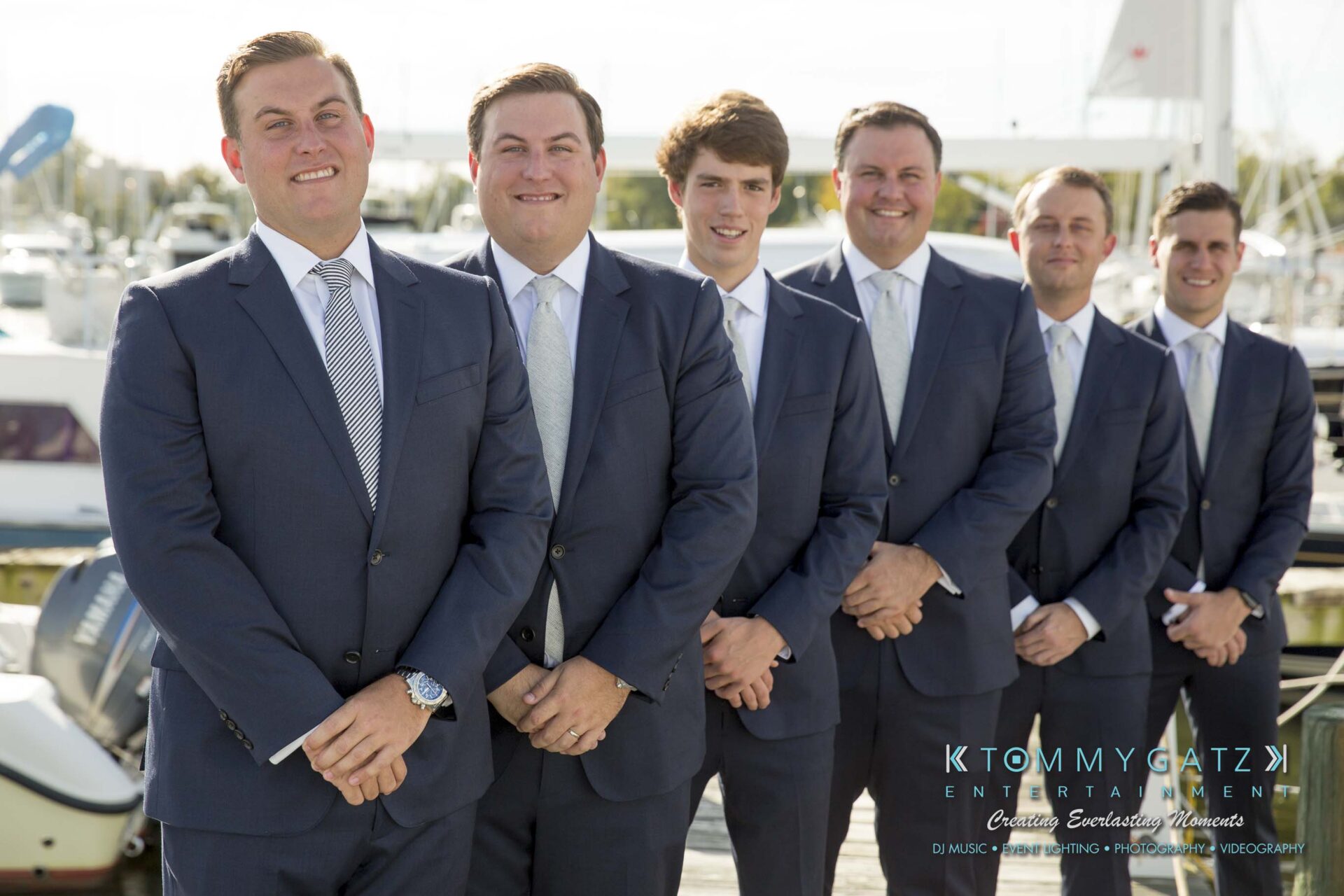 Six men in suits stand in a row, smiling, with boats and water in the background.