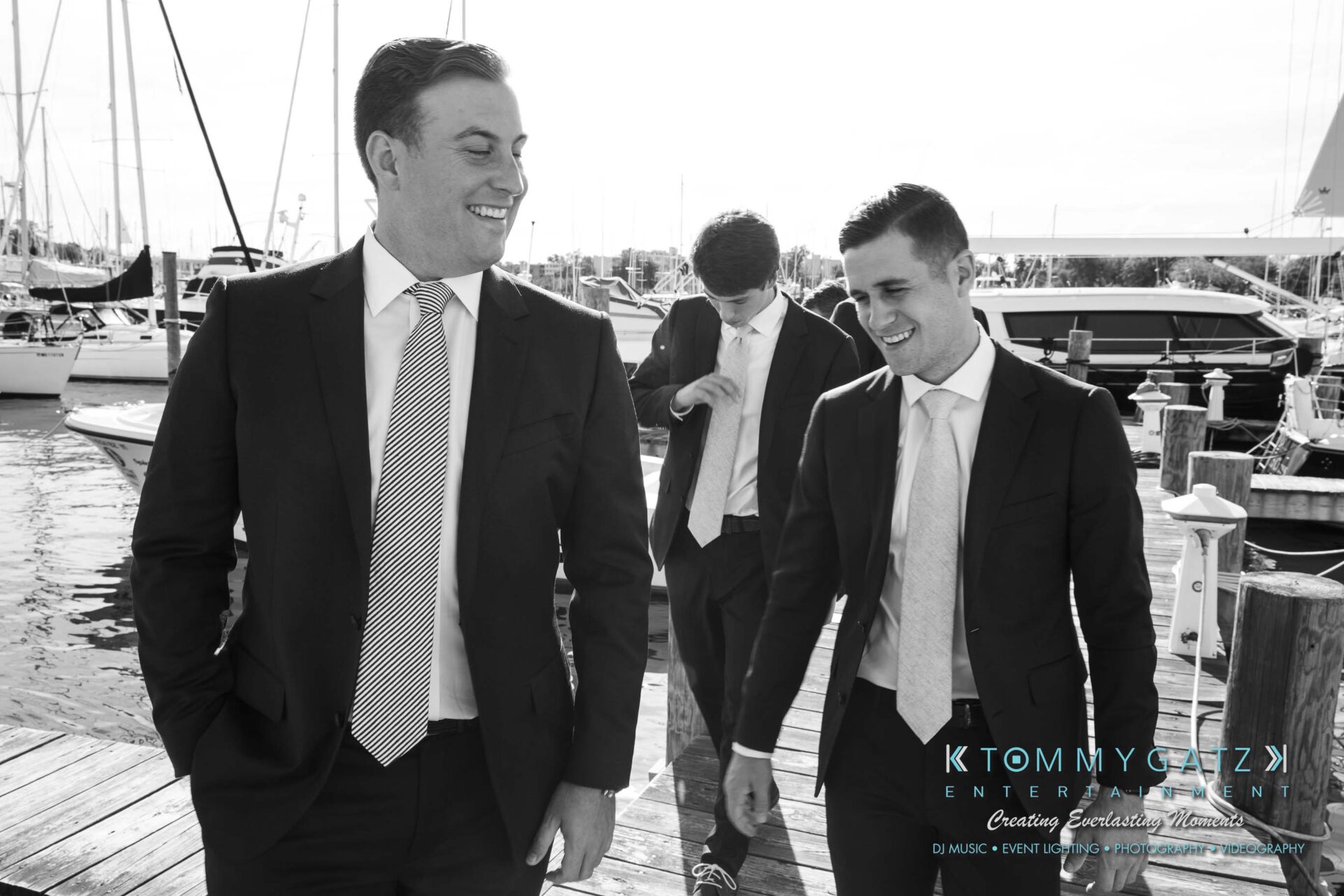Amanda-Tyler-102117-PREP-Low-Res-WM-108 - TOMMY GATZ ENTERTAINMENT Three men in suits walk and talk on a dock by a marina with boats in the background.