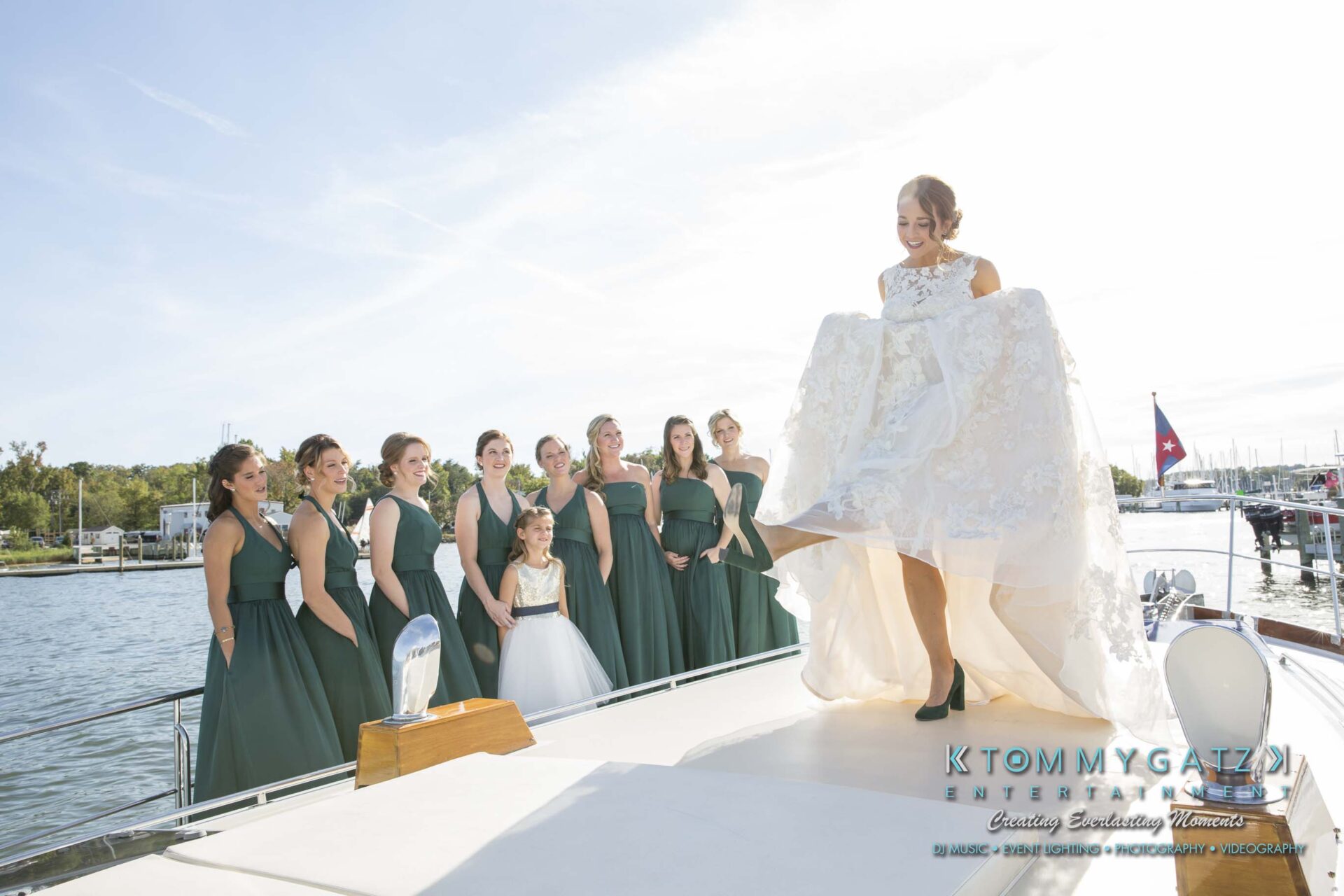 Amanda-Tyler-102117-PREP-Low-Res-WM-112 - TOMMY GATZ ENTERTAINMENT A bride lifts her dress while standing on a boat, surrounded by bridesmaids in green dresses and a girl in white, against a backdrop of water and trees.