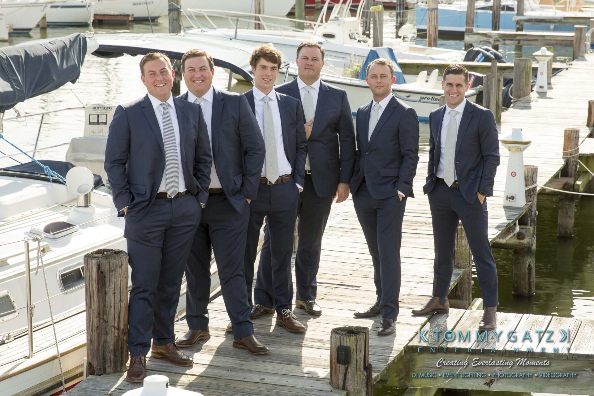 Amanda-Tyler-102117-PREP-Low-Res-WM-114 - TOMMY GATZ ENTERTAINMENT Six men in matching suits stand on a wooden dock with boats in the background, posing for a group photo.