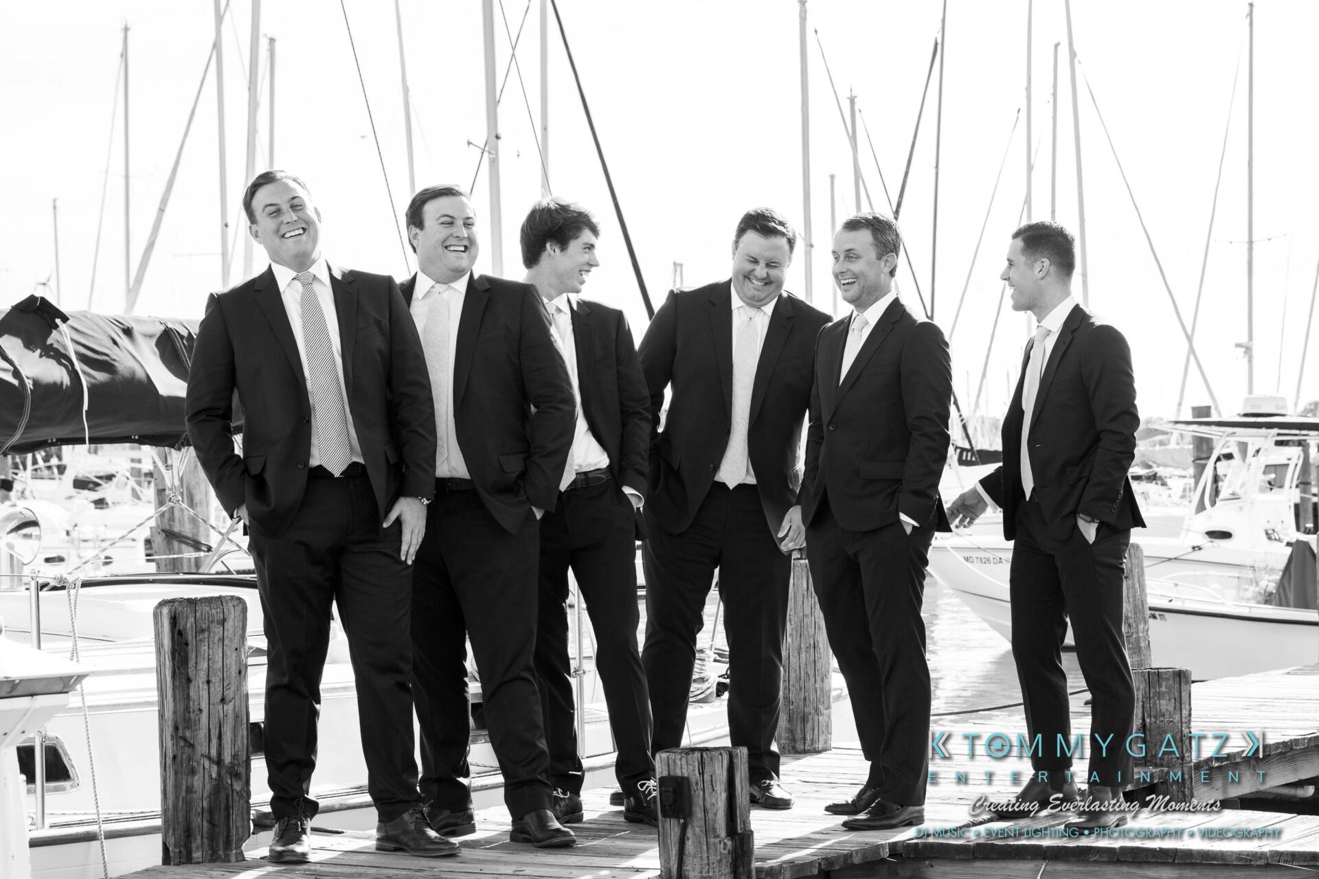 Amanda-Tyler-102117-PREP-Low-Res-WM-115 - TOMMY GATZ ENTERTAINMENT Six men in suits stand and laugh on a dock with boats in the background.