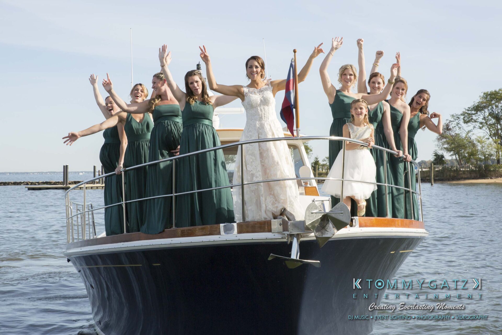 A bride and bridesmaids in green dresses stand on a boat, raising their arms. It's a sunny day by the water.