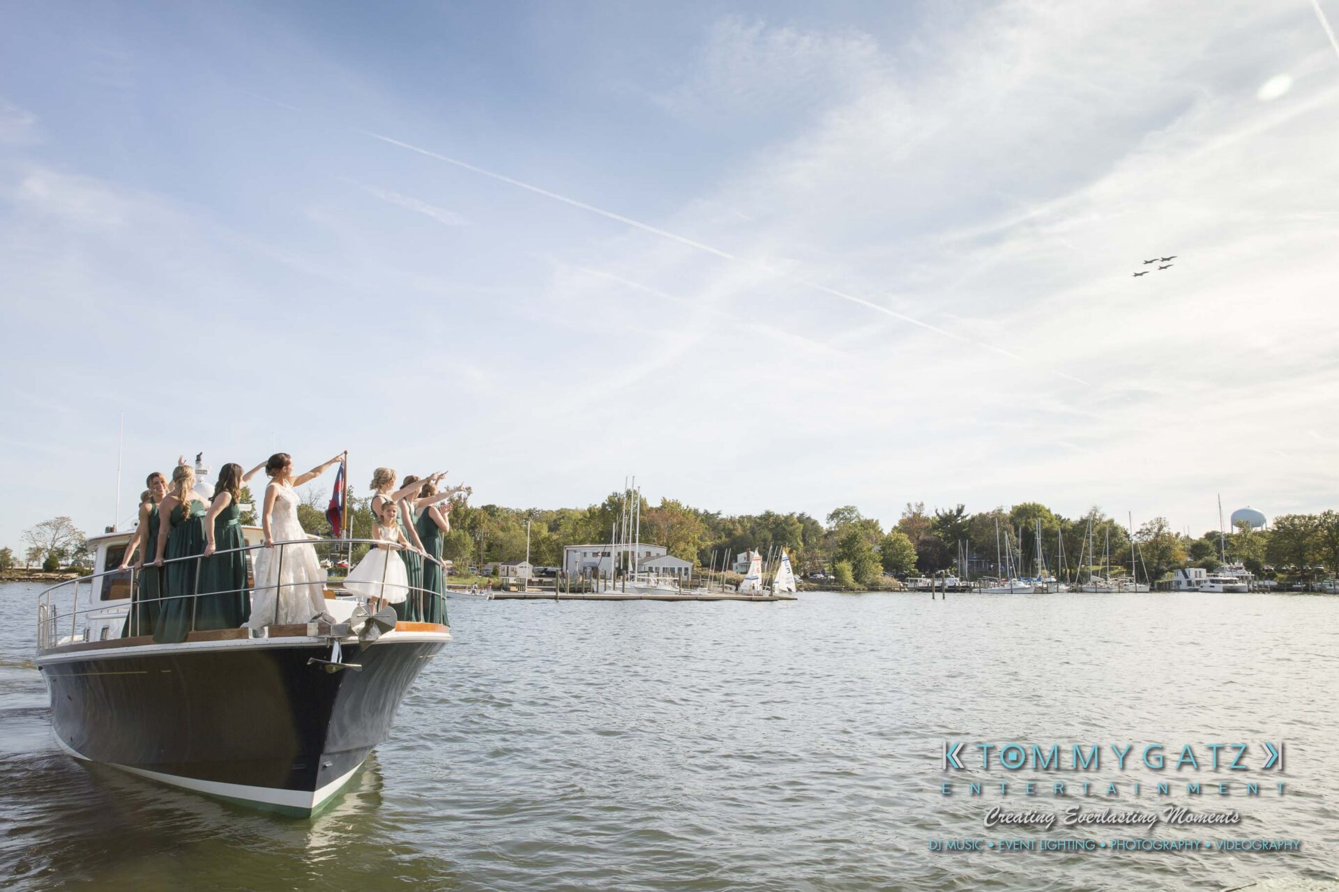 Amanda-Tyler-102117-PREP-Low-Res-WM-132 - TOMMY GATZ ENTERTAINMENT A group of people in formal attire wave from the bow of a boat on a sunny day, with a marina and trees in the background and planes flying overhead.