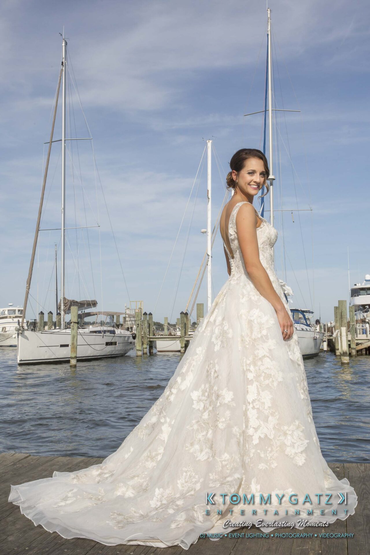 A bride in a lace wedding dress stands on a dock with boats and water in the background.