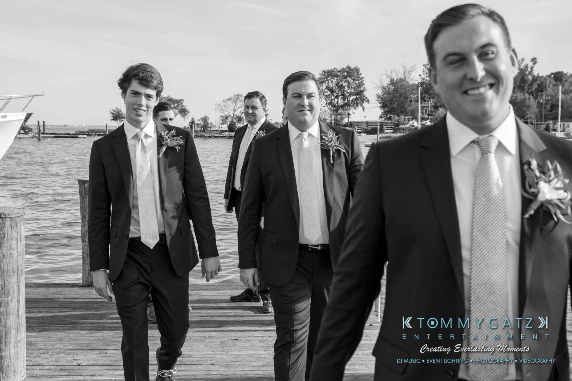 Three men in suits walking on a wooden dock by the water, smiling and wearing boutonnieres.