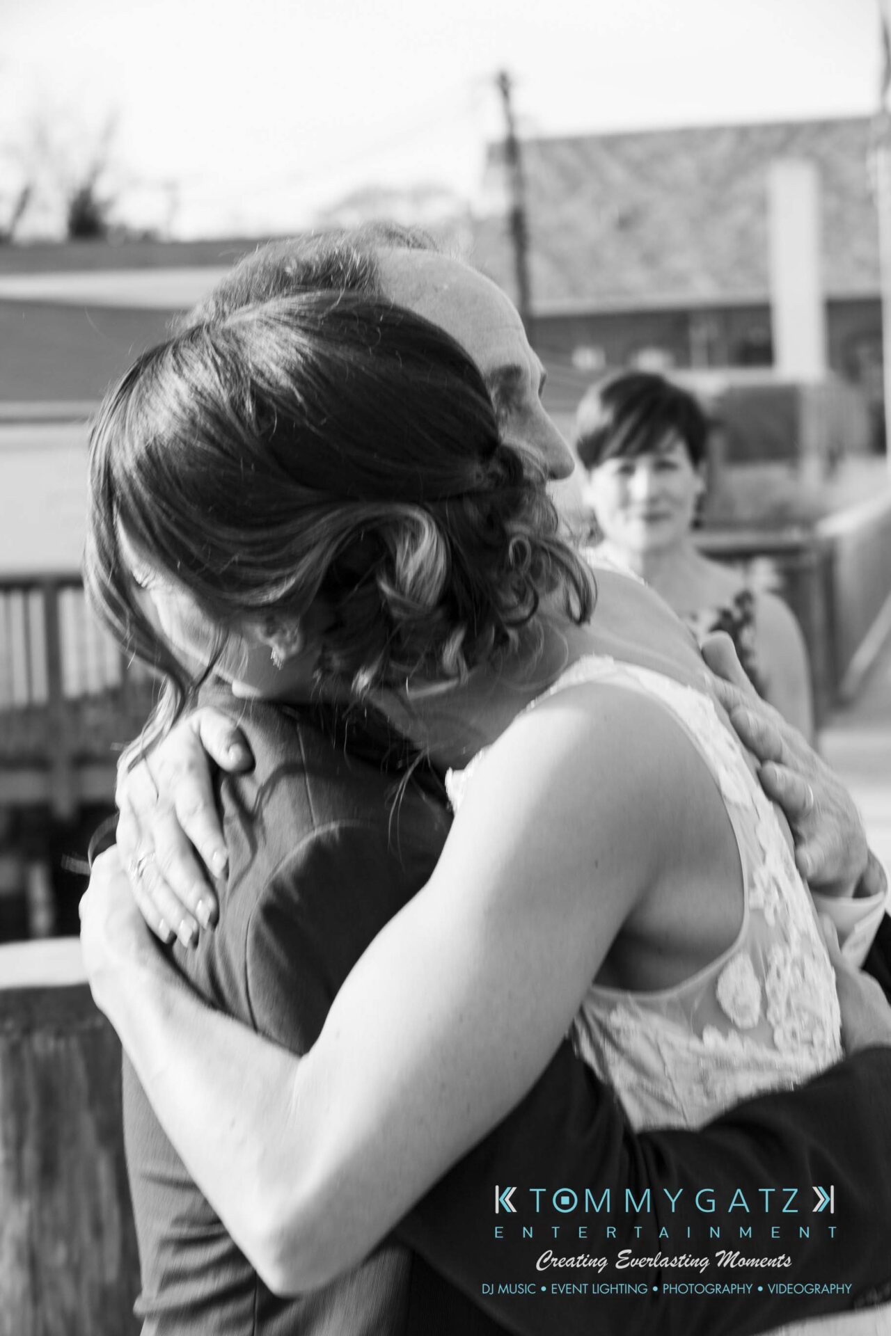 A woman in a white dress hugs a man in an embrace. A person in the background watches. There is a logo for a business in the bottom corner.