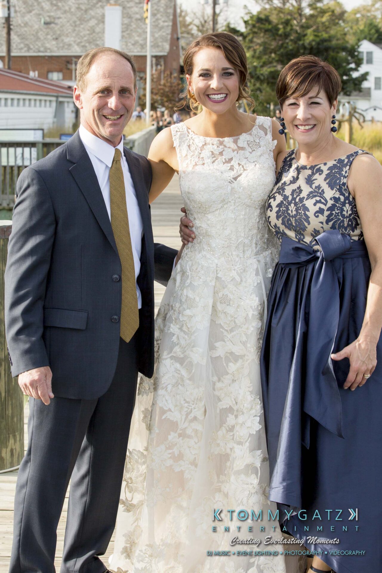 Amanda-Tyler-102117-PREP-Low-Res-WM-181-1 - TOMMY GATZ ENTERTAINMENT Three people posing outdoors; a man in a suit, a woman in a white lace dress, and another woman in a floral gown. They are standing on a wooden walkway near buildings.