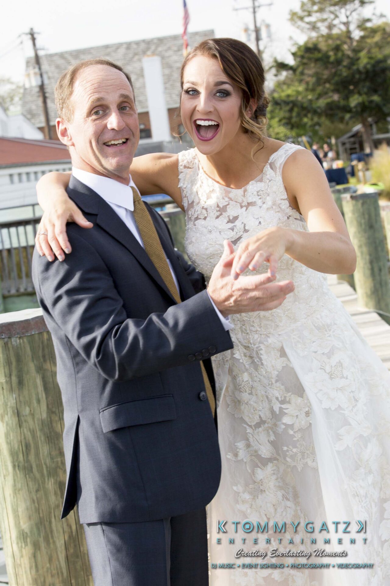A bride in a white dress joyfully poses with a man in a suit outdoors on a sunny day.