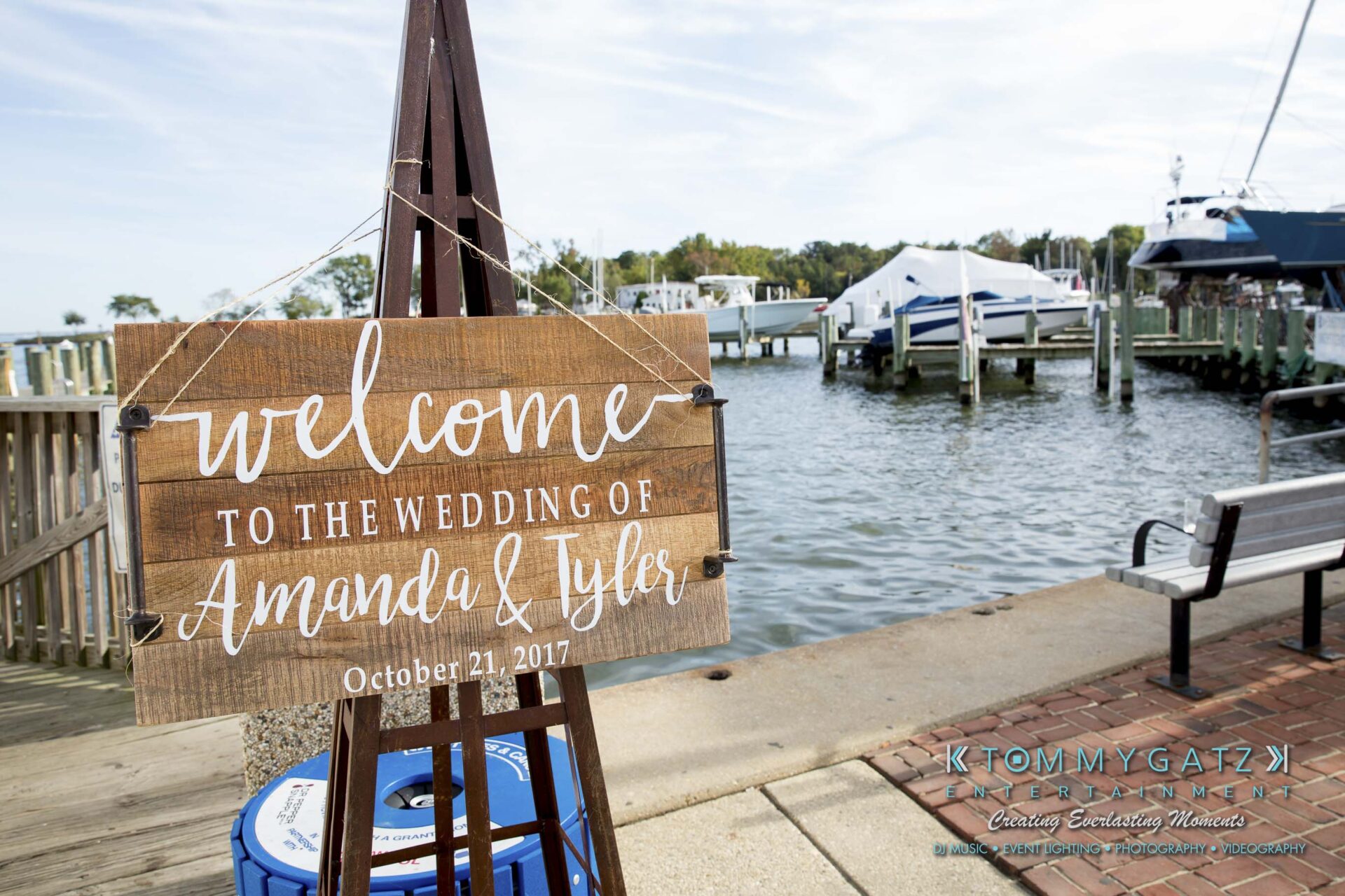 A wooden sign reads: "Welcome to the wedding of Amanda & Tyler, October 21, 2017." It stands by a marina with boats and water in the background.