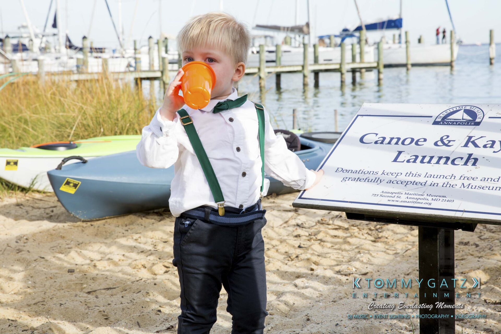 Amanda-Tyler-102117-PREP-Low-Res-WM-198 - TOMMY GATZ ENTERTAINMENT Young child in formal attire drinks from an orange cup beside a canoe and kayak launch sign, with boats and water in the background.