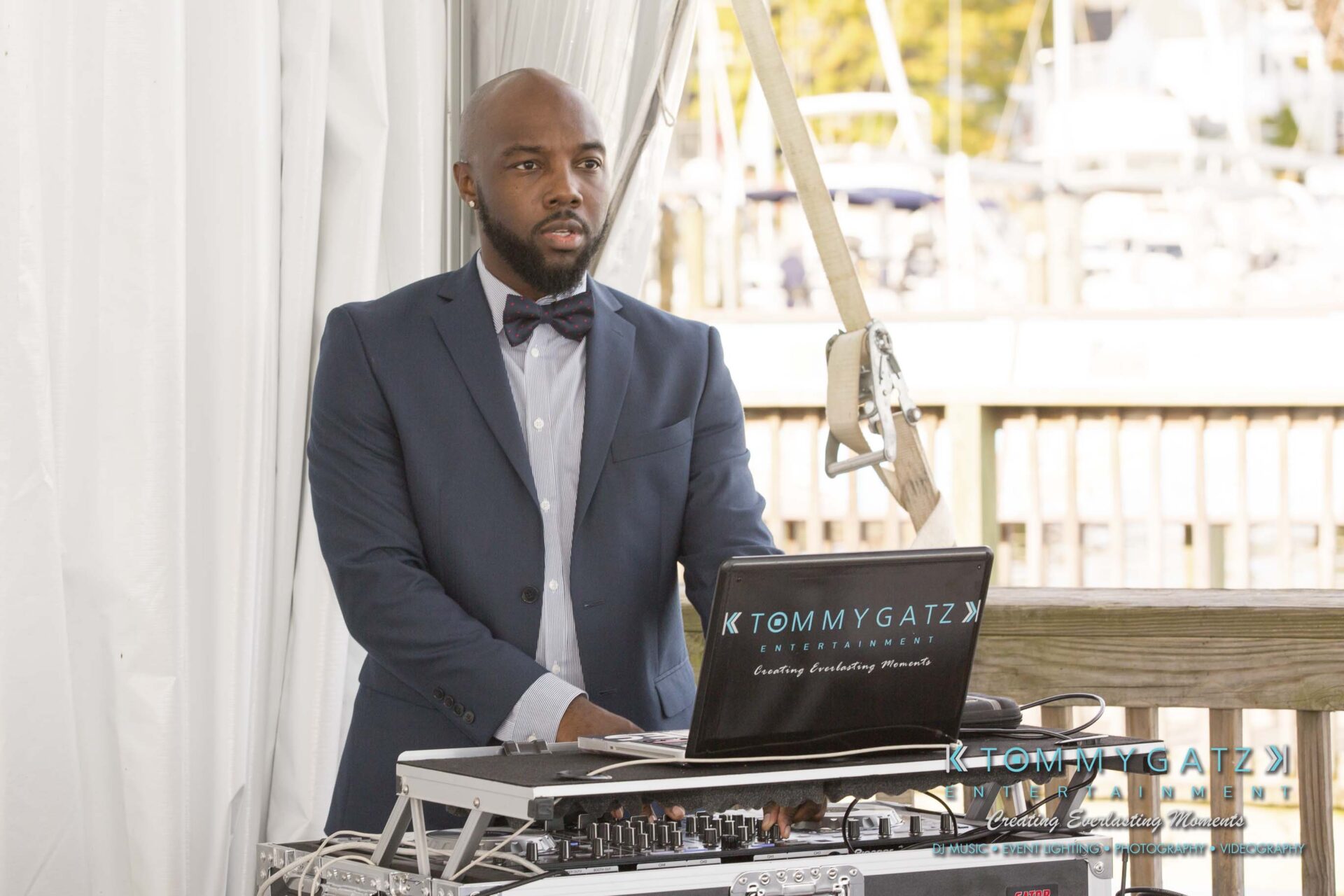 A man in a suit stands behind DJ equipment outdoors, with "Tommy Gatz Entertainment" displayed on the laptop.