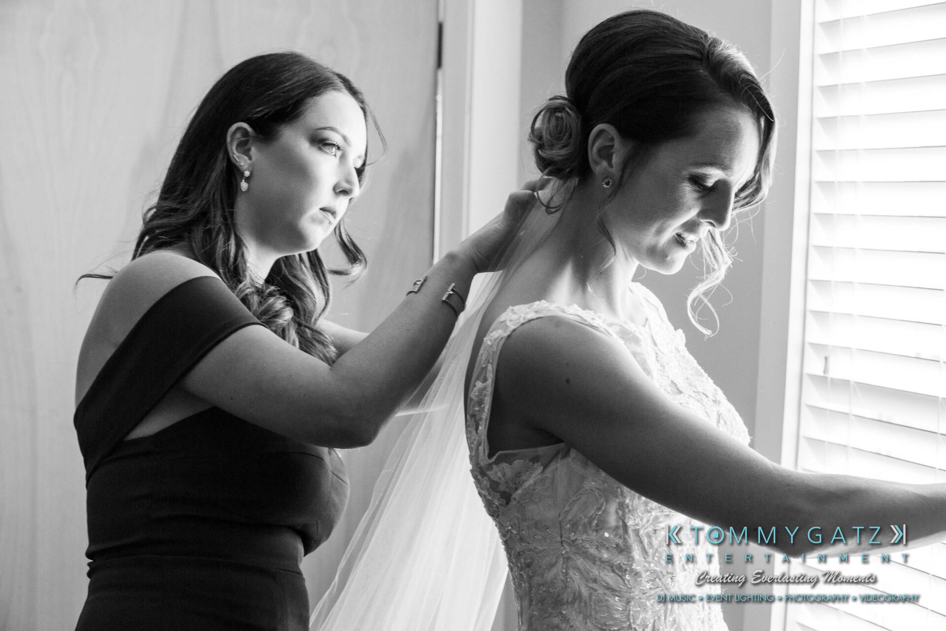 A woman in a black dress helps a bride adjust her veil near a window.