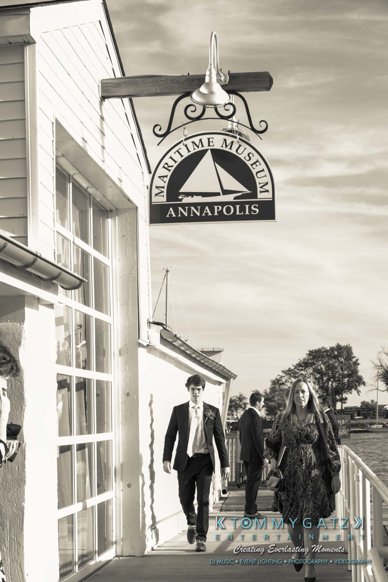 People walking on a boardwalk near the Annapolis Maritime Museum, with a lamp above displaying the museum's name.