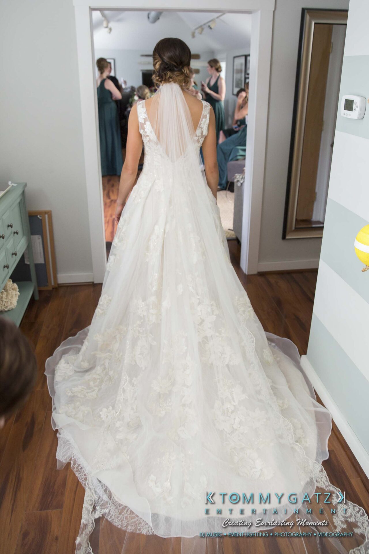 Amanda-Tyler-102117-PREP-Low-Res-WM-219 - TOMMY GATZ ENTERTAINMENT A bride in a white wedding dress with a long train is standing in a room. Bridesmaids in dark dresses are visible in the background.