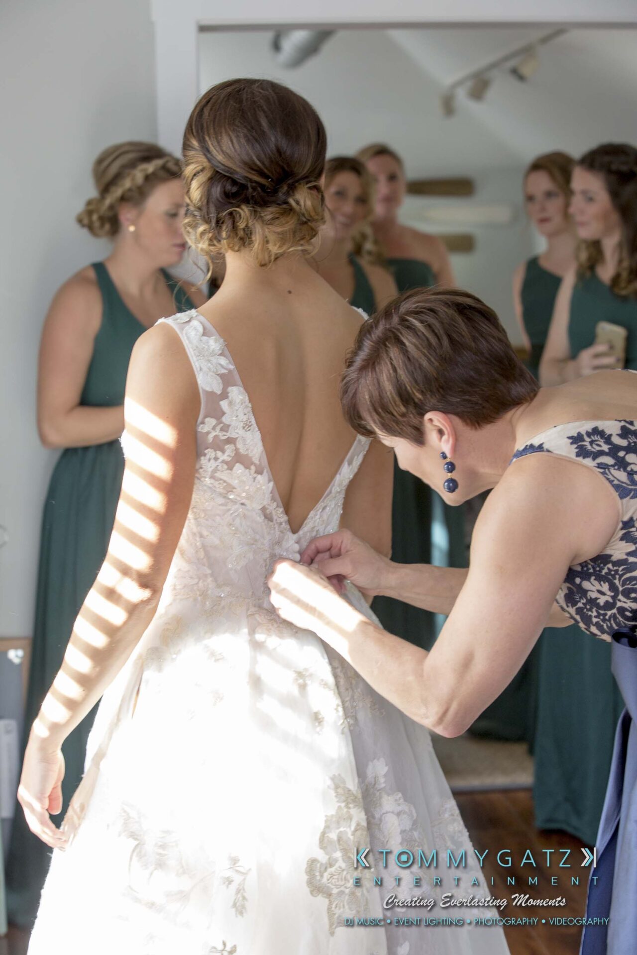 A woman fastens the back of a bride's lace wedding dress while bridesmaids in green dresses look on.
