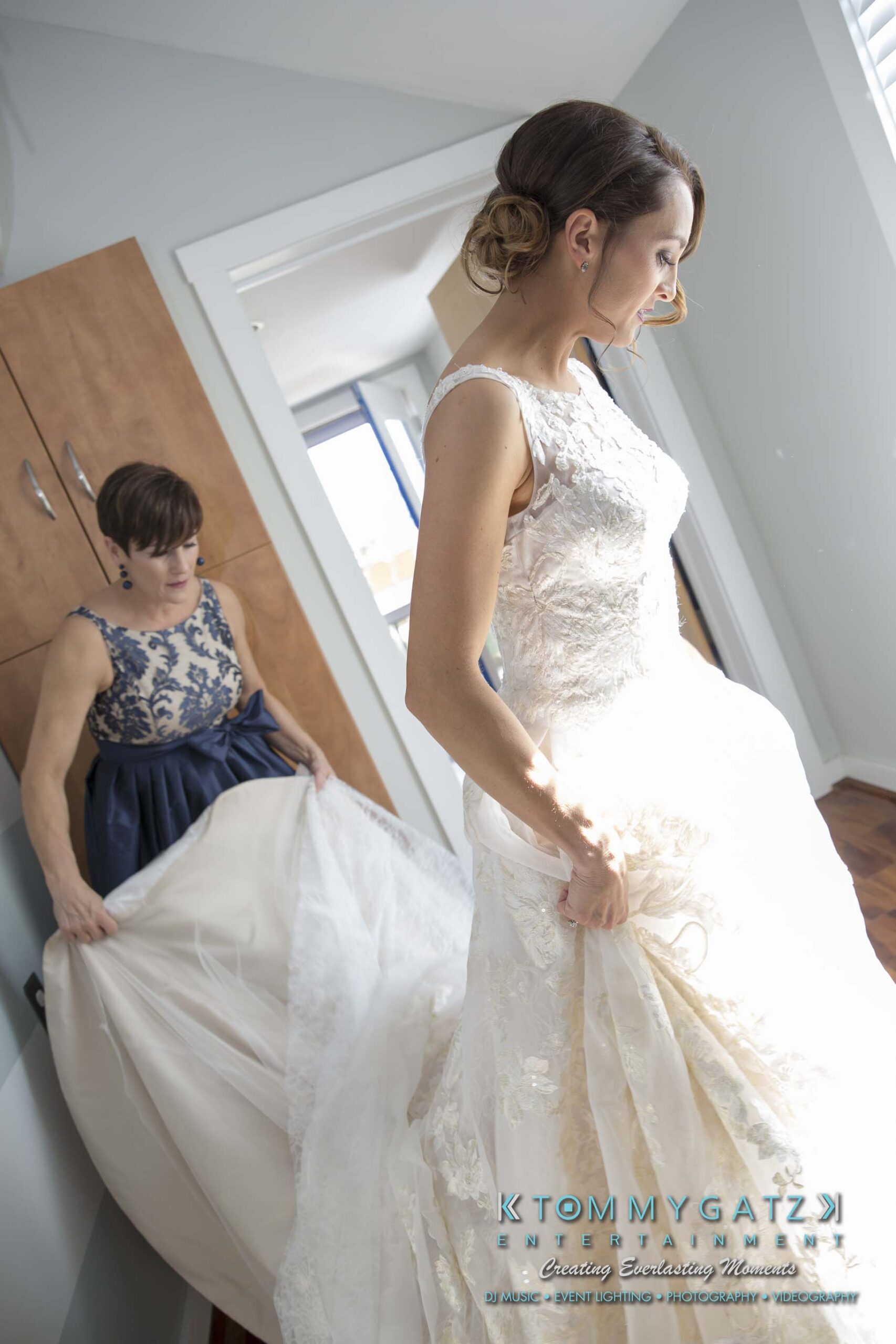 Bride in white dress stands in a room adjusting her gown, while another woman in a blue dress helps with the train.
