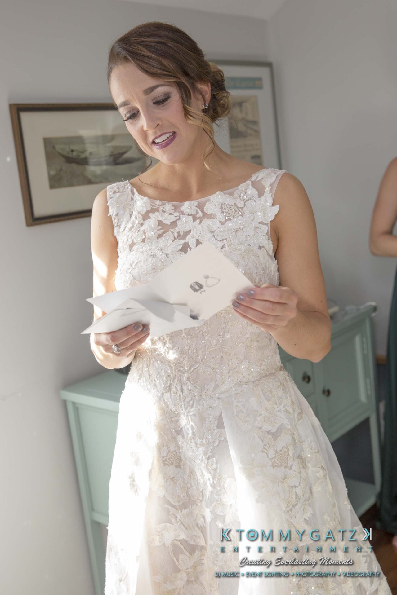 Amanda-Tyler-102117-PREP-Low-Res-WM-36 - TOMMY GATZ ENTERTAINMENT A woman in a white lace dress reads a card, standing indoors near a light blue cabinet.
