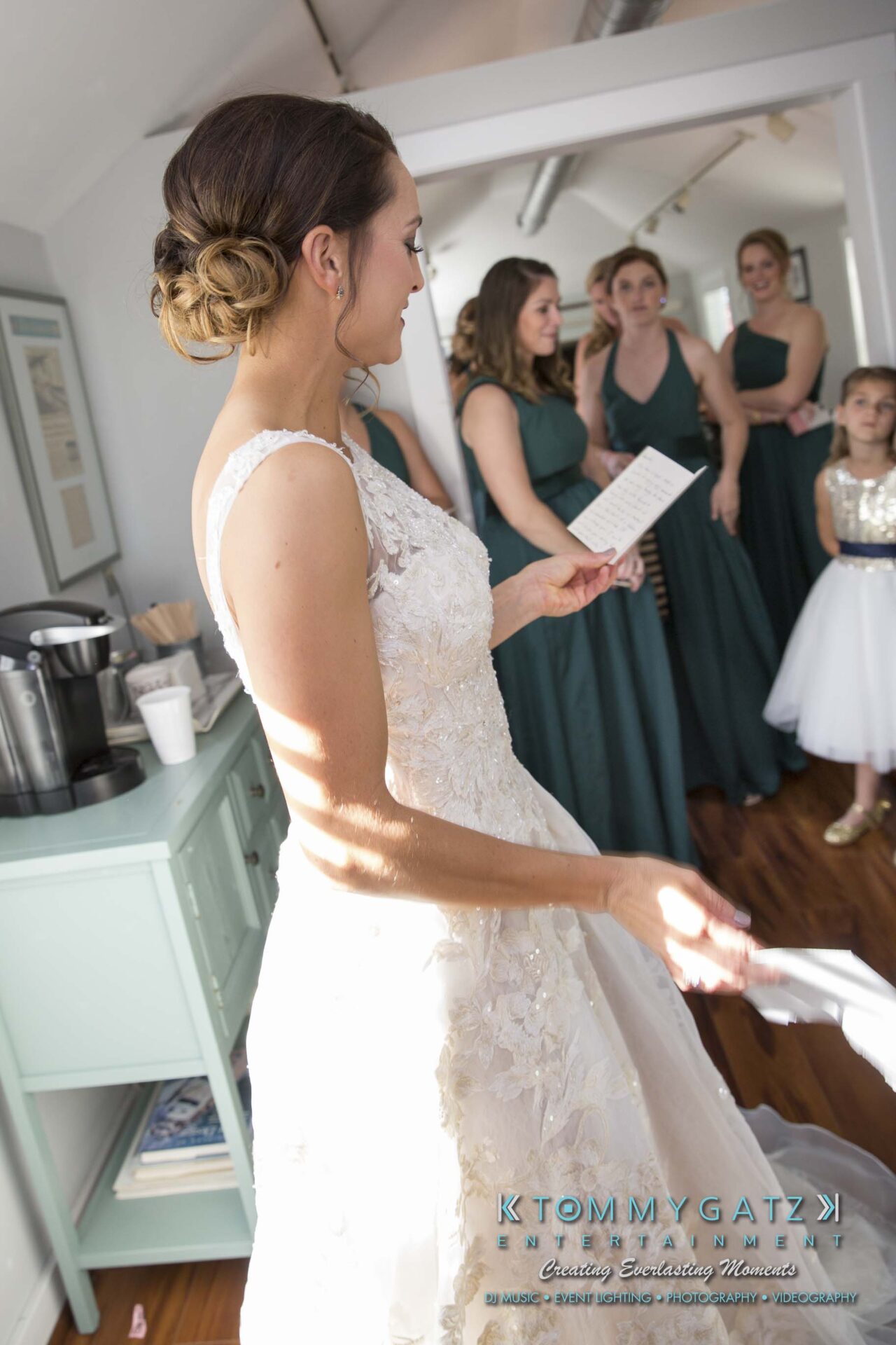 Amanda-Tyler-102117-PREP-Low-Res-WM-39 - TOMMY GATZ ENTERTAINMENT Bride in white gown holds a paper, surrounded by bridesmaids in green dresses and a young girl in white, in a bright room.