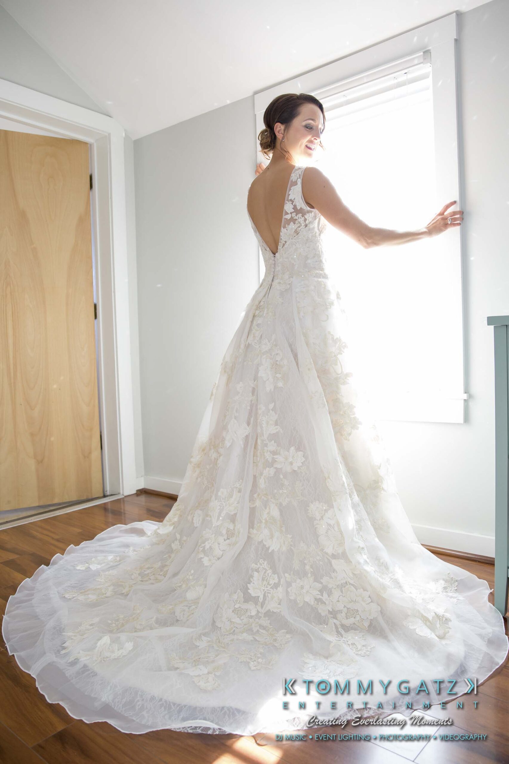 Amanda-Tyler-102117-PREP-Low-Res-WM-54 - TOMMY GATZ ENTERTAINMENT Bride in a white lace wedding dress with a long train, standing near a window, smiling over her shoulder.