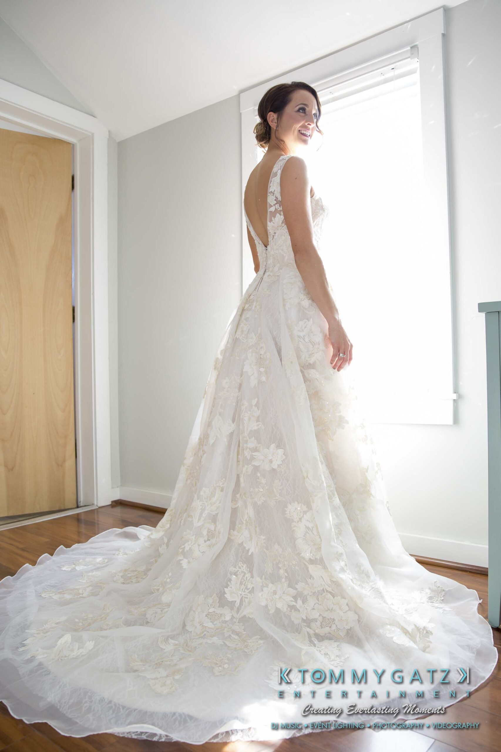 A bride in a white floral gown stands inside, smiling and looking over her shoulder near a window.