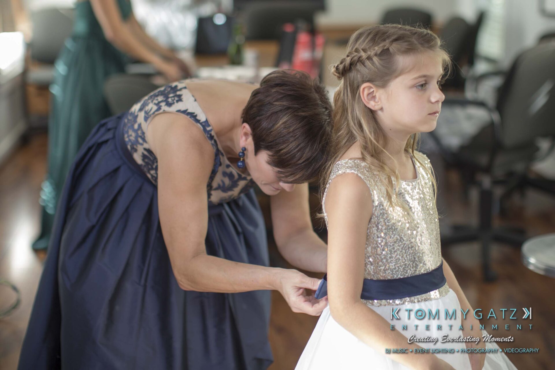 Amanda-Tyler-102117-PREP-Low-Res-WM-6 - TOMMY GATZ ENTERTAINMENT A woman in a blue dress helps a young girl in a white and silver dress adjust her belt in a well-lit room.
