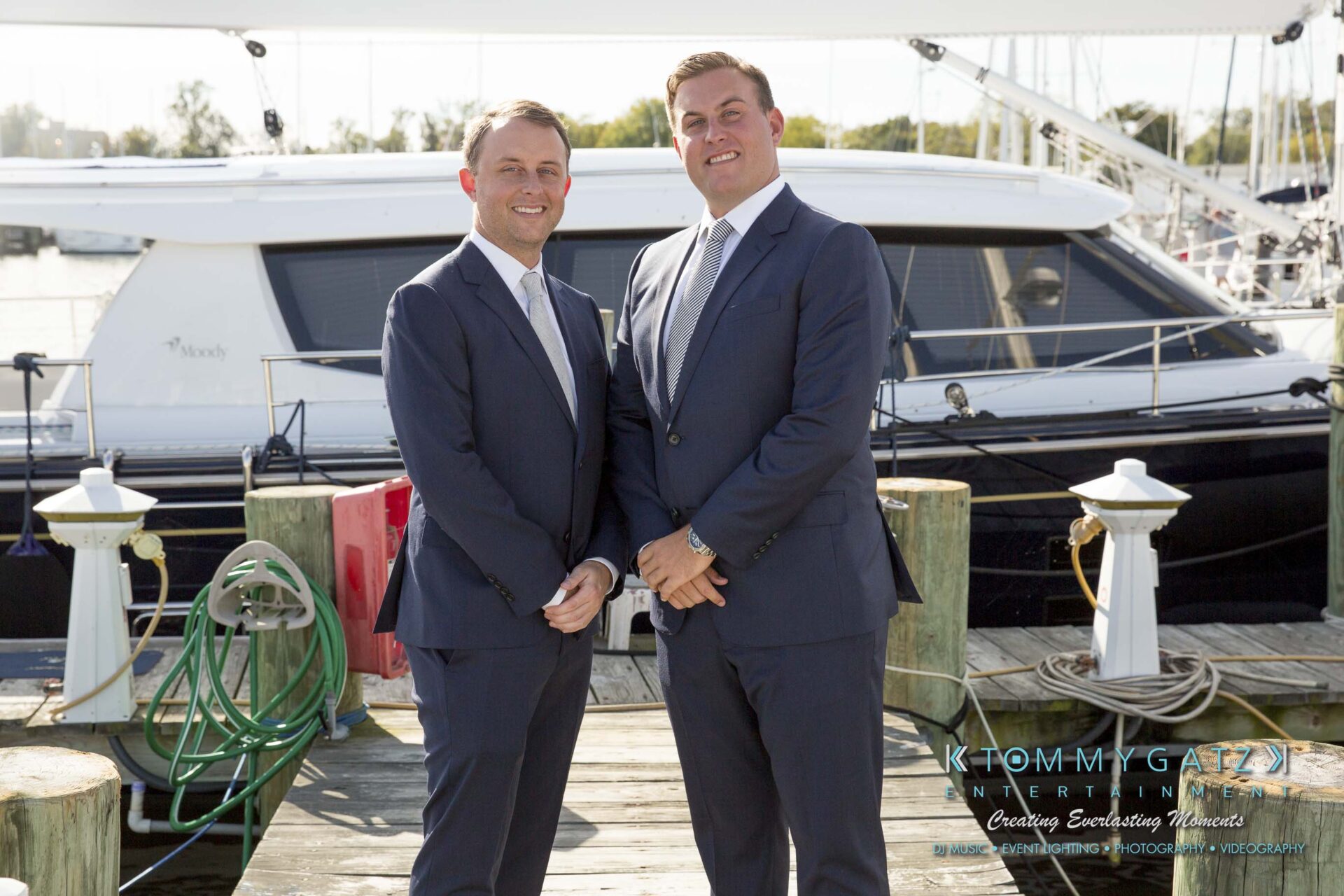 Amanda-Tyler-102117-PREP-Low-Res-WM-75 - TOMMY GATZ ENTERTAINMENT Two men in suits stand on a dock in front of a large yacht, with ropes and boats visible in the background.