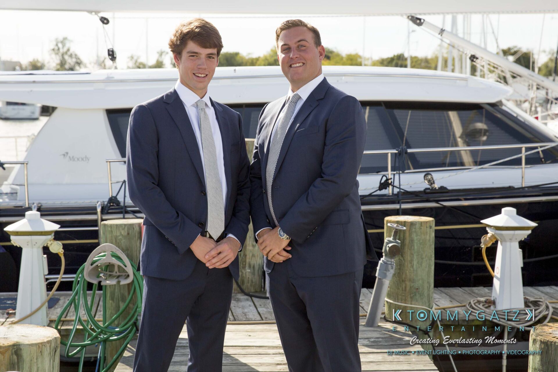Amanda-Tyler-102117-PREP-Low-Res-WM-80 - TOMMY GATZ ENTERTAINMENT Two men in suits stand on a dock with boats in the background, smiling at the camera.