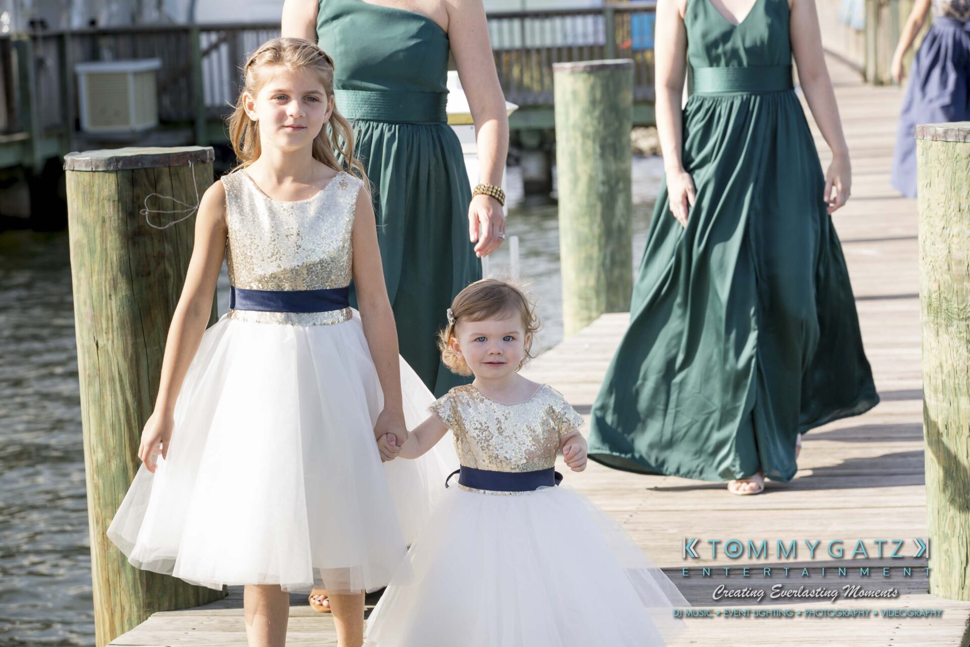 Amanda-Tyler-102117-PREP-Low-Res-WM-84 - TOMMY GATZ ENTERTAINMENT Two young girls in white dresses walk hand in hand on a wooden dock, followed by women in green dresses.