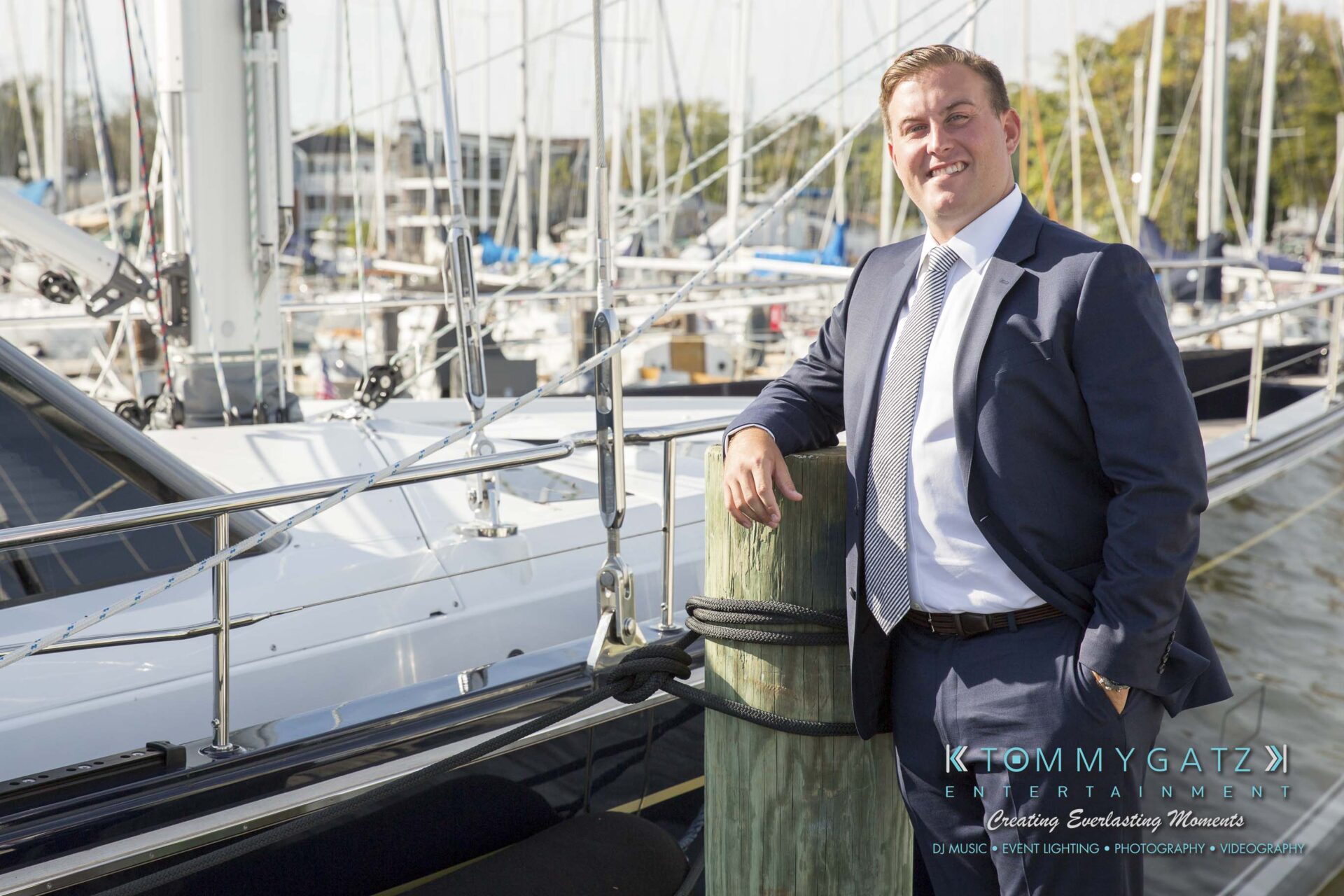Amanda-Tyler-102117-PREP-Low-Res-WM-88 - TOMMY GATZ ENTERTAINMENT A man in a suit stands by a yacht, smiling with one arm resting on a dock post. Sailboats are in the background.