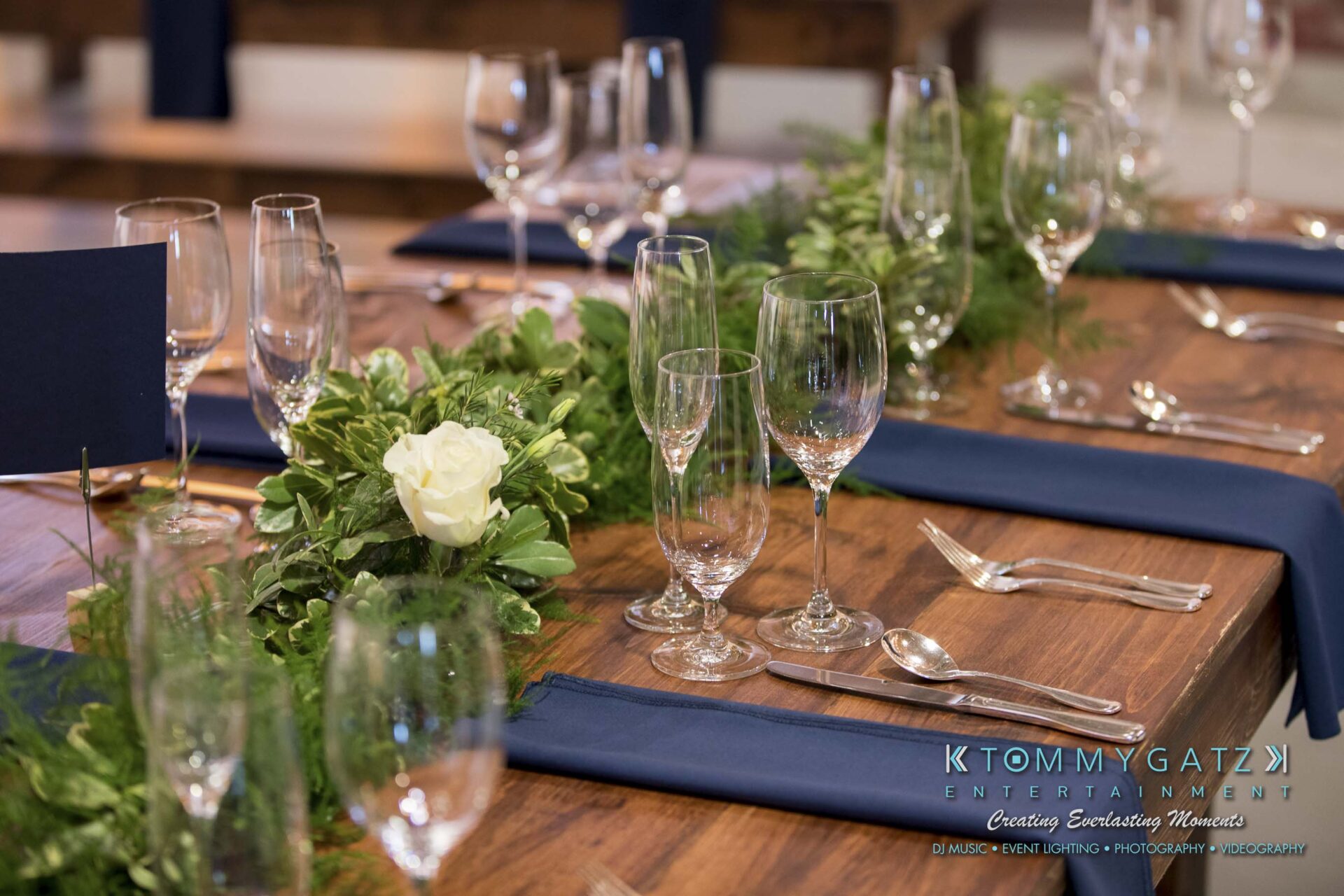 Amanda-Tyler-102117-RECEPTION-Low-Res-WM-1 - TOMMY GATZ ENTERTAINMENT A table is set with empty wine glasses, cutlery, navy blue napkins, and green floral centerpieces with white roses.