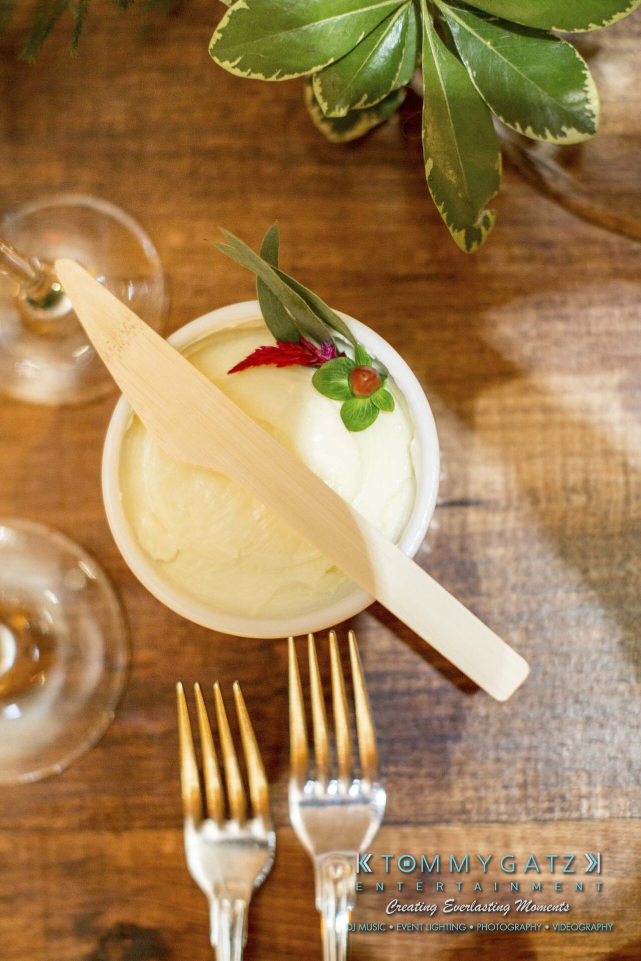 Amanda-Tyler-102117-RECEPTION-Low-Res-WM-11 - TOMMY GATZ ENTERTAINMENT A bowl of creamy dessert garnished with green leaves and a pink flower, topped with a wooden utensil. Nearby are a wine glass and three forks on a wooden table with leafy decor.