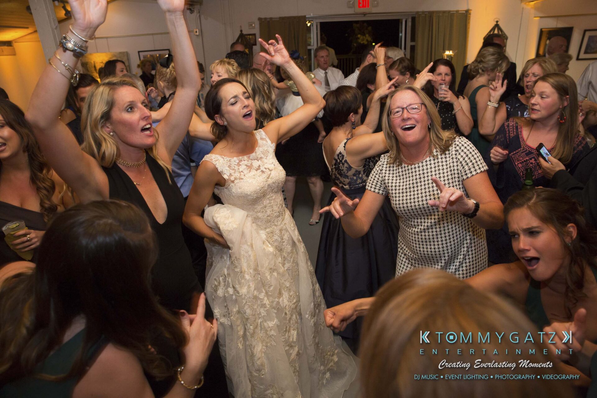 Amanda-Tyler-102117-RECEPTION-Low-Res-WM-116 - TOMMY GATZ ENTERTAINMENT A bride in a lace gown dances energetically with guests on a crowded dance floor, surrounded by people raising their hands and singing.