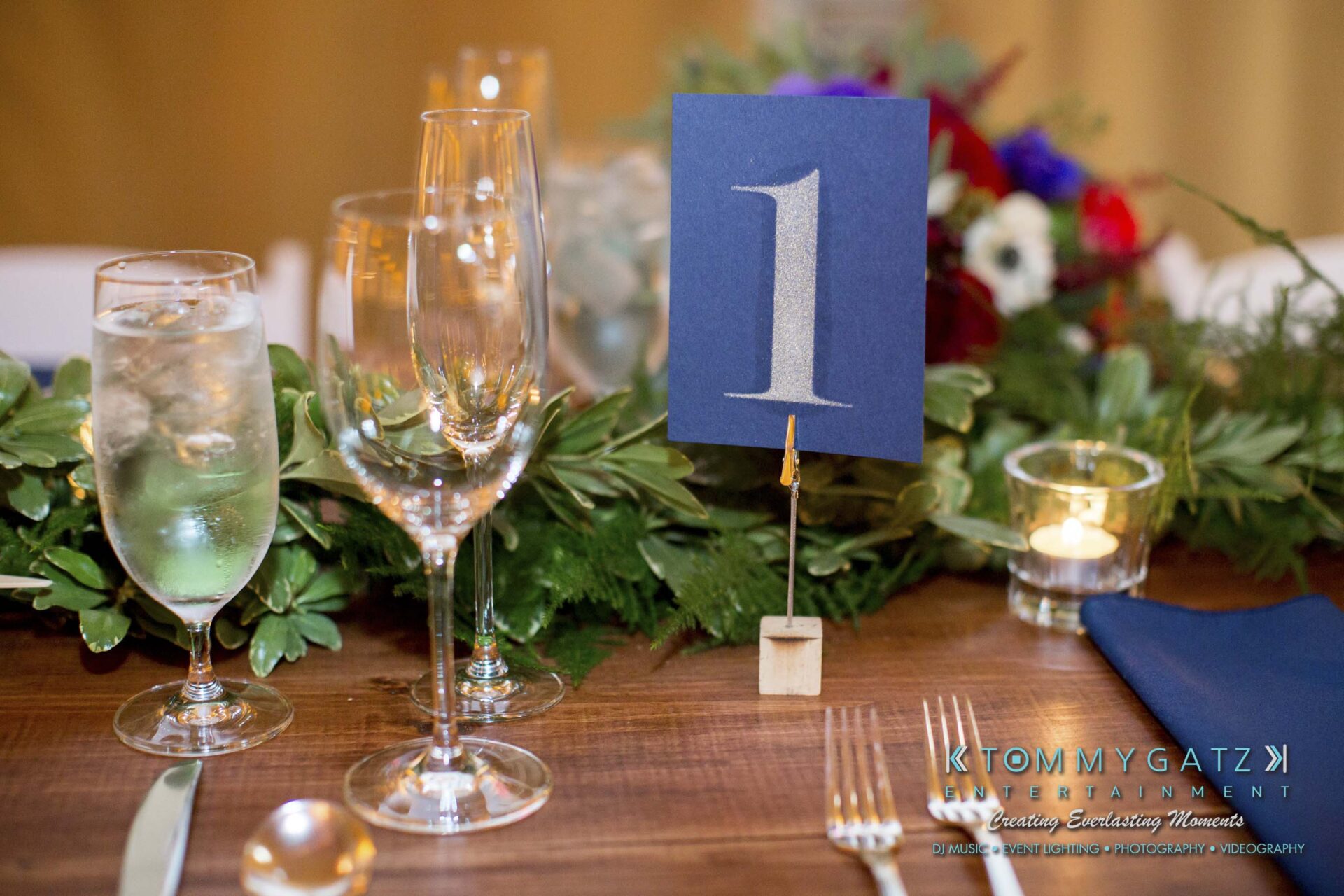 Amanda-Tyler-102117-RECEPTION-Low-Res-WM-12 - TOMMY GATZ ENTERTAINMENT Elegant table setting with wine glasses, a water glass, cutlery, and a table number "1" sign. Green foliage and small candles are used as decoration.