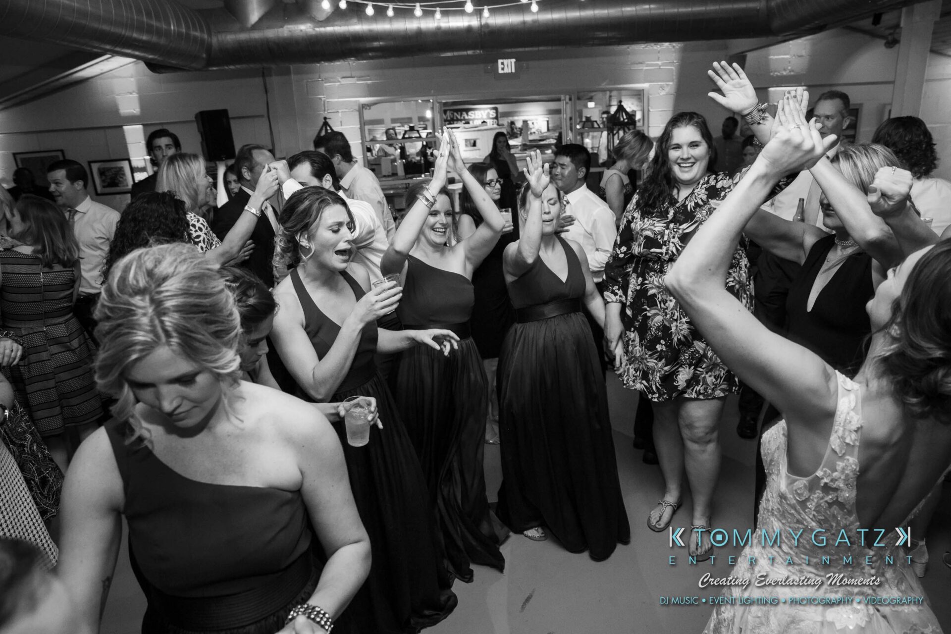 Amanda-Tyler-102117-RECEPTION-Low-Res-WM-123 - TOMMY GATZ ENTERTAINMENT A lively group of people dancing and enjoying an indoor event, with women in dresses raising their arms and others holding drinks.