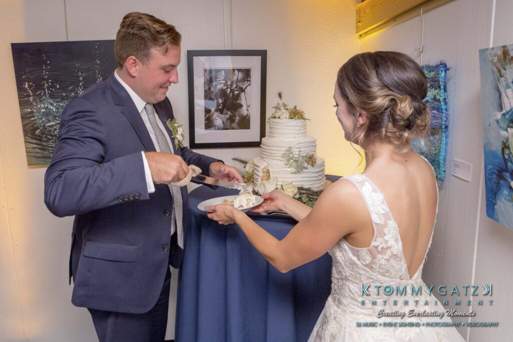 Amanda-Tyler-102117-RECEPTION-Low-Res-WM-154 - TOMMY GATZ ENTERTAINMENT A bride and groom are cutting a multi-tiered wedding cake together, with the groom holding a cake slice and the bride holding a plate.