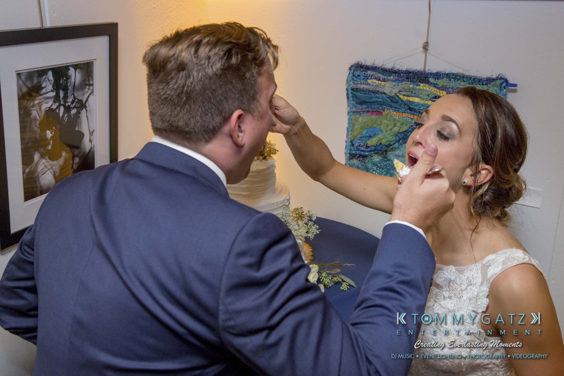Amanda-Tyler-102117-RECEPTION-Low-Res-WM-155 - TOMMY GATZ ENTERTAINMENT A bride and groom feed each other cake at a wedding reception, with a framed photo and a tapestry on the wall behind them.
