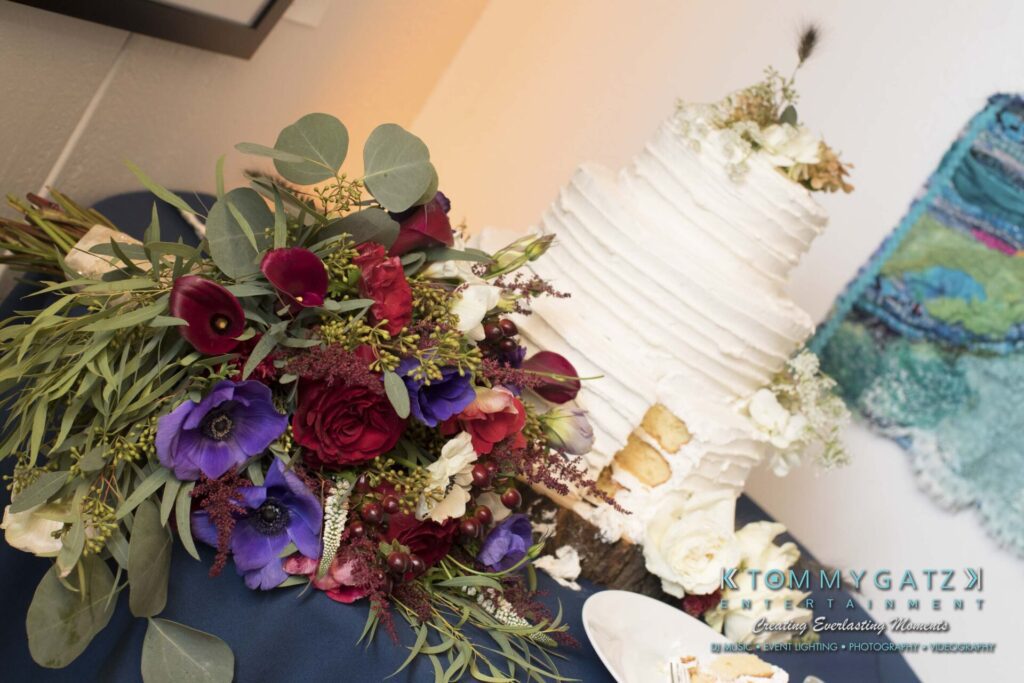 Amanda-Tyler-102117-RECEPTION-Low-Res-WM-158 - TOMMY GATZ ENTERTAINMENT A three-tiered white cake with floral decoration sits beside a large bouquet of mixed flowers and greenery on a dark cloth.