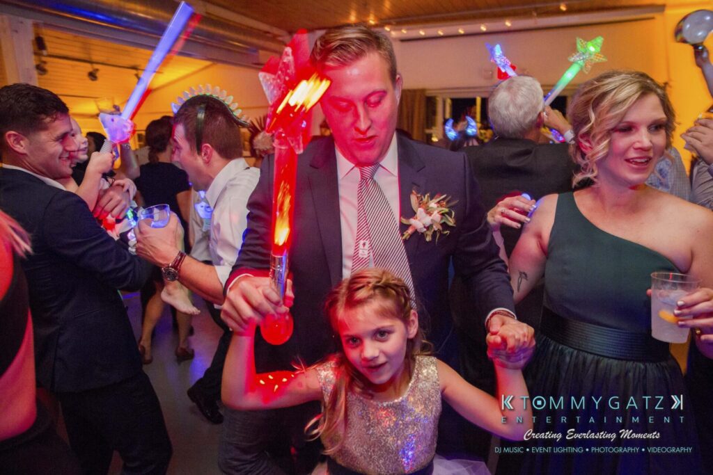 A man and a girl holding light-up toys dance at a party, surrounded by other people. Bright lights and festive decorations are visible.