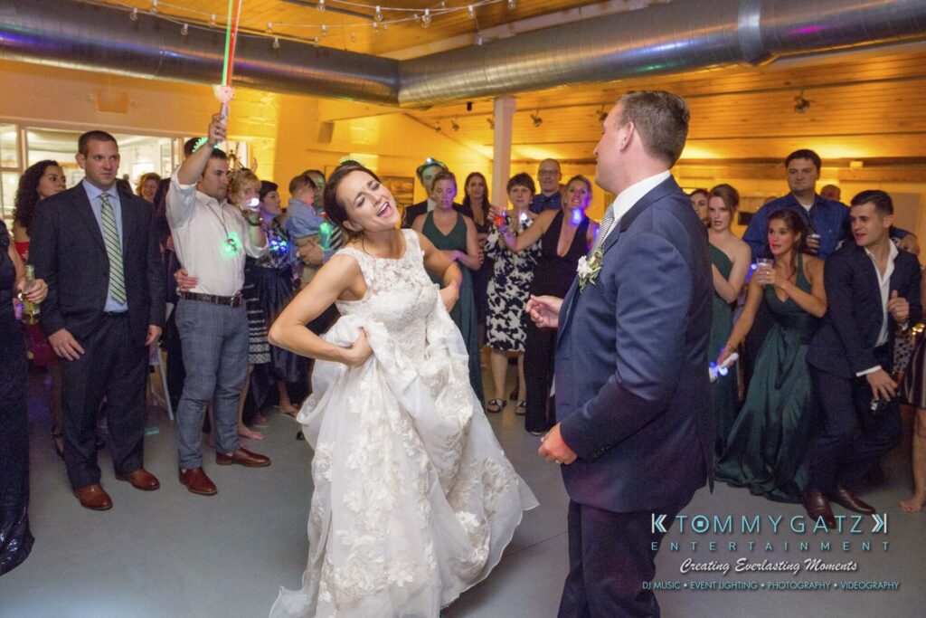 A bride and groom dance enthusiastically at their wedding reception, surrounded by guests in a warmly lit venue.