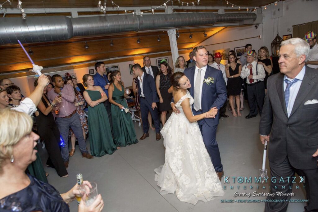 A bride and groom dance at a wedding reception, surrounded by guests. A woman in the foreground holds a light stick.